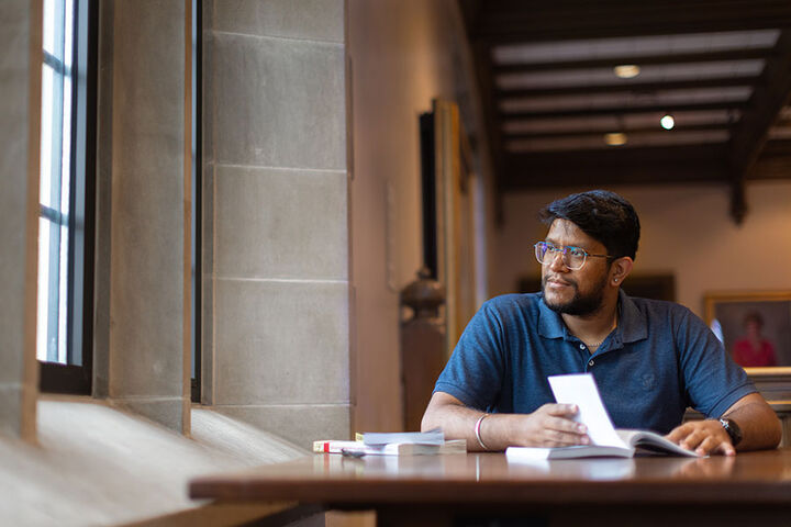 A male student looks out a window while sitting at a table with books.